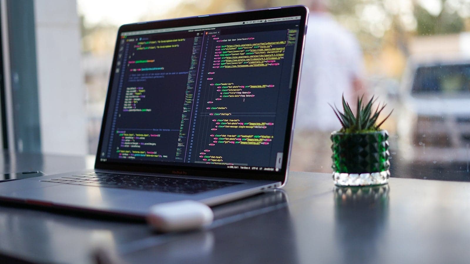 窓辺の机に置かれた、コードを表示したノート PC と小さな多肉植物